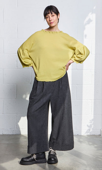 Plümo- Woman in a Pansy Blouse and wide-leg dark gray pants stands against a white brick wall, hands in pockets.