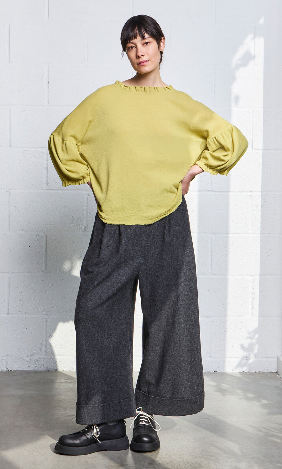 Plümo- Woman in a Pansy Blouse and wide-leg dark gray pants stands against a white brick wall, hands in pockets.