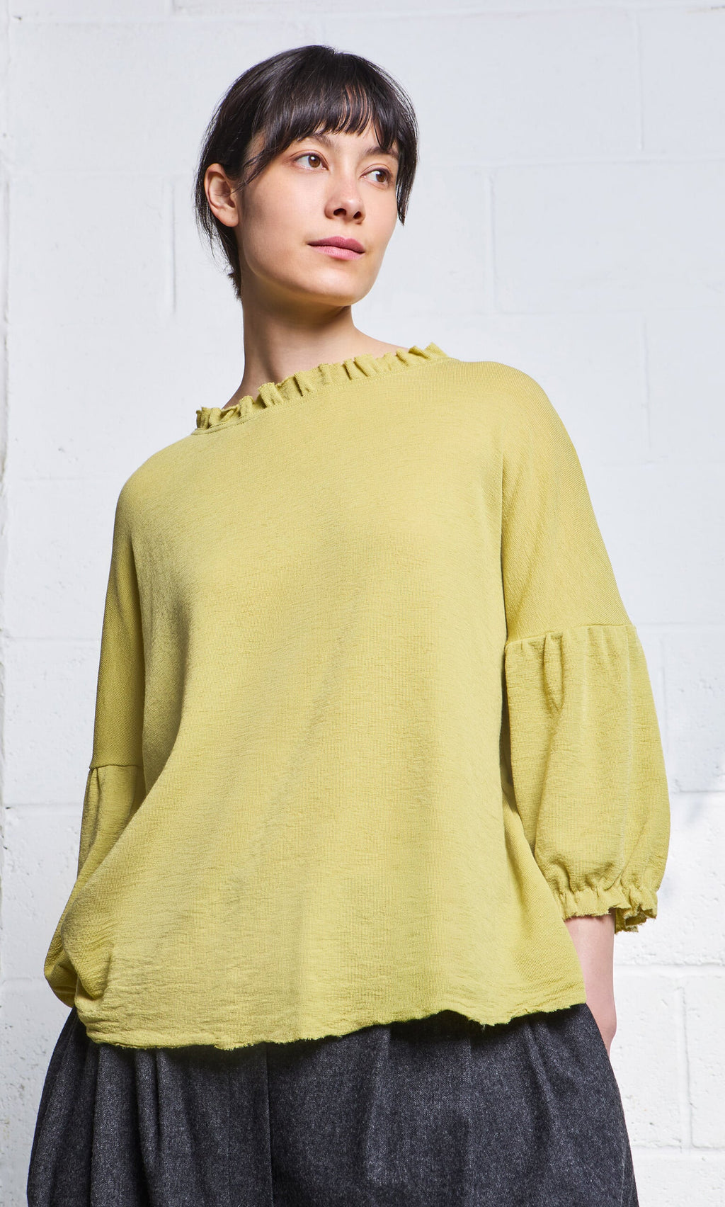 Plümo- Woman wearing the Pansy Blouse in light green, standing against a white brick wall.