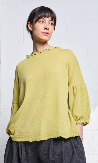 Plümo- Woman wearing the Pansy Blouse in light green, standing against a white brick wall.