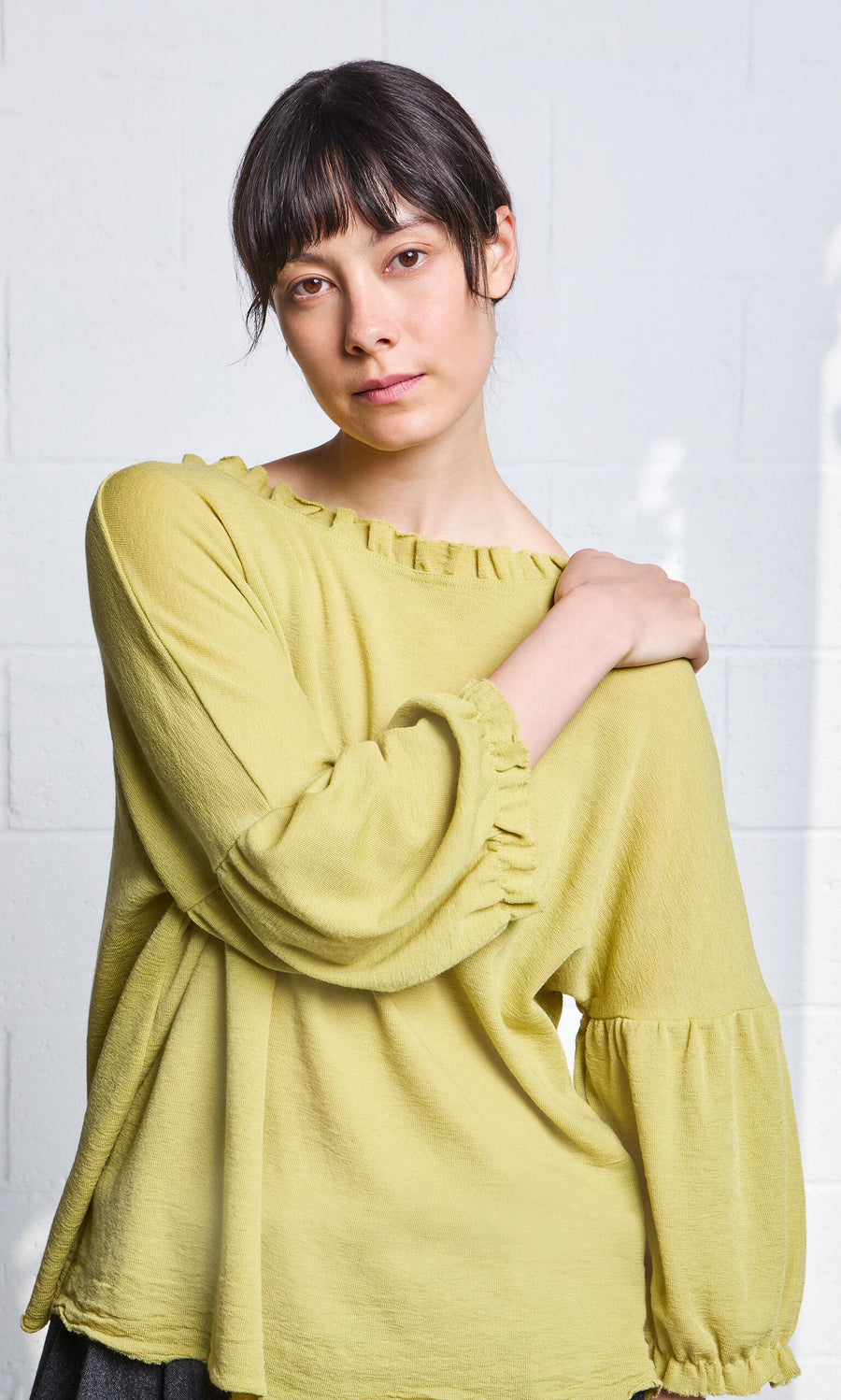 Plümo- Woman with short dark hair wears the Pansy Blouse, posing against a light brick wall background.