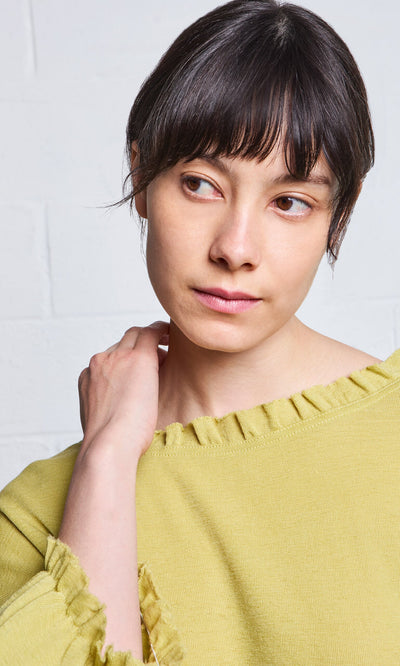 Plümo- A woman with short dark hair wears the Pansy Blouse, looking to the side and touching her neck with a neutral expression.