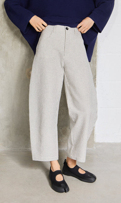 Plümo- Person wearing lightweight Passanti Pants, a navy top, and black tabi shoes, standing against a concrete wall.