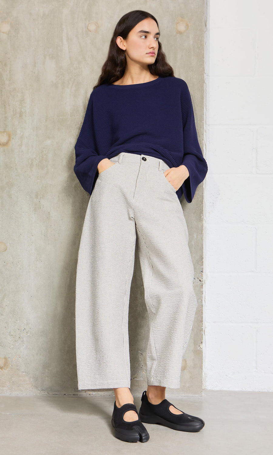 Plümo- Woman in a navy top and Passanti Pants stands against a concrete wall, wearing black shoes.