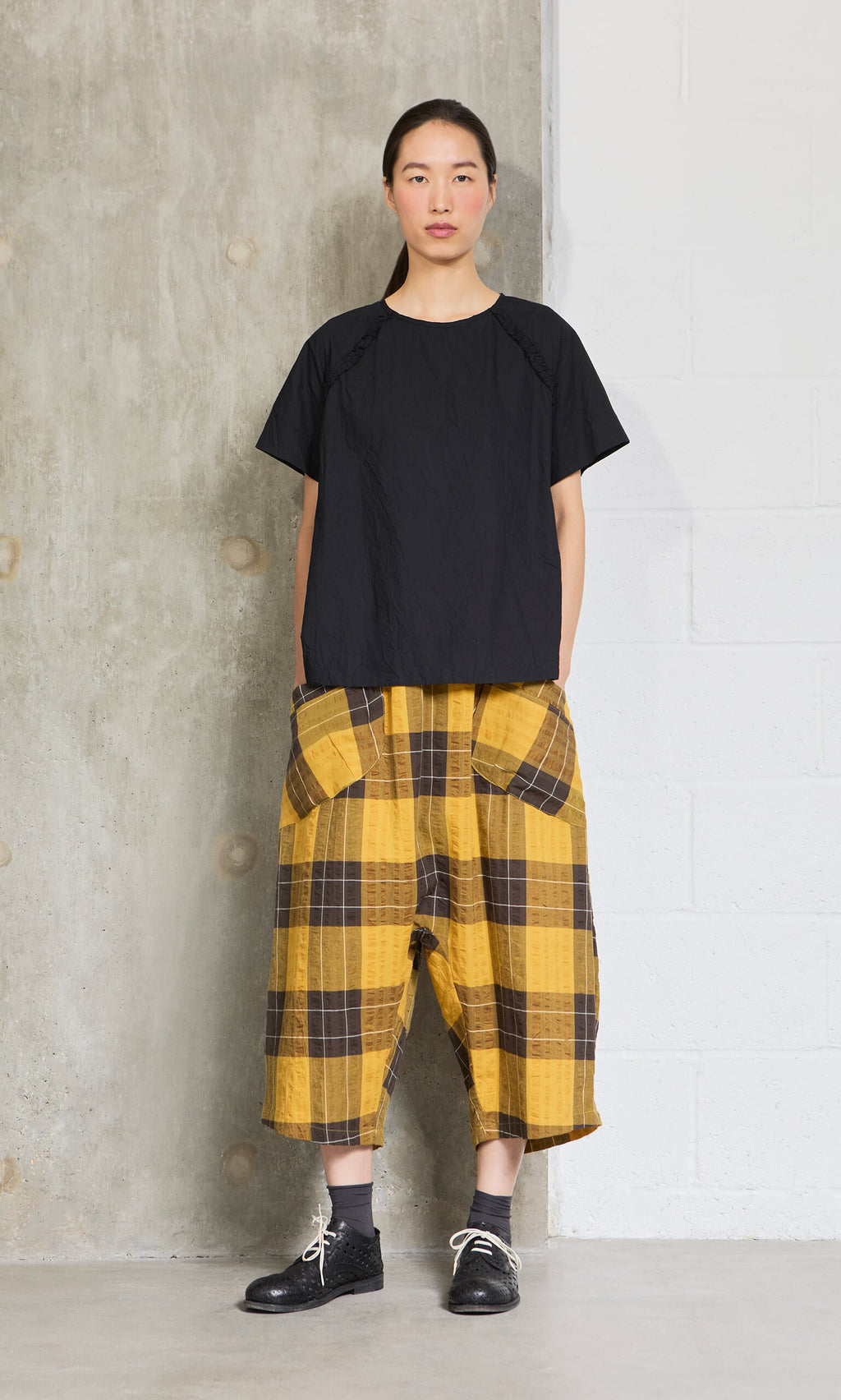 Plümo- Woman in a crinkly black blouse and yellow plaid wide-leg pants with large pockets, standing by a concrete wall.
