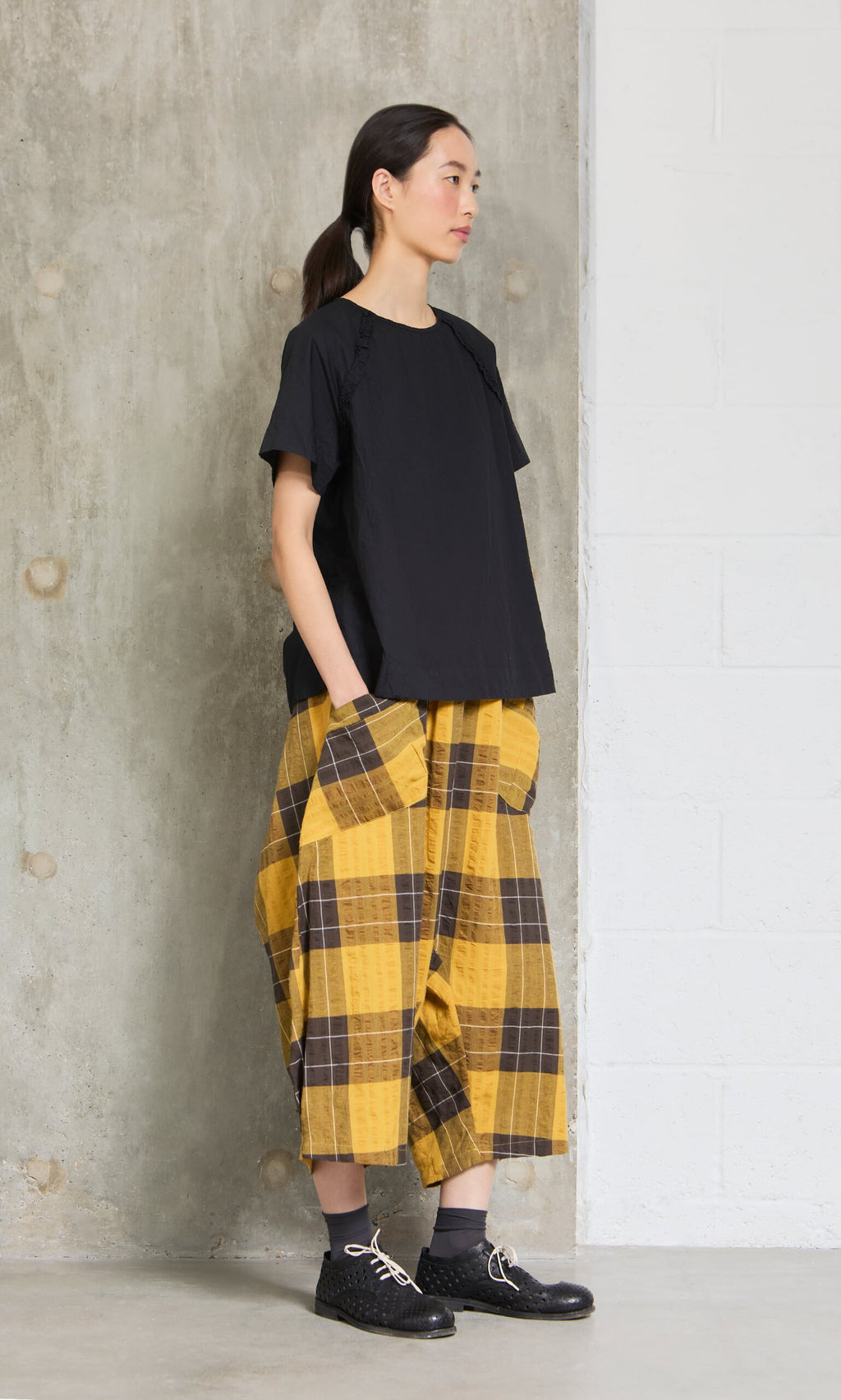 Plümo- Woman in a Crinkly Black Blouse and yellow plaid wide-leg pants with large pockets, standing by a concrete wall.