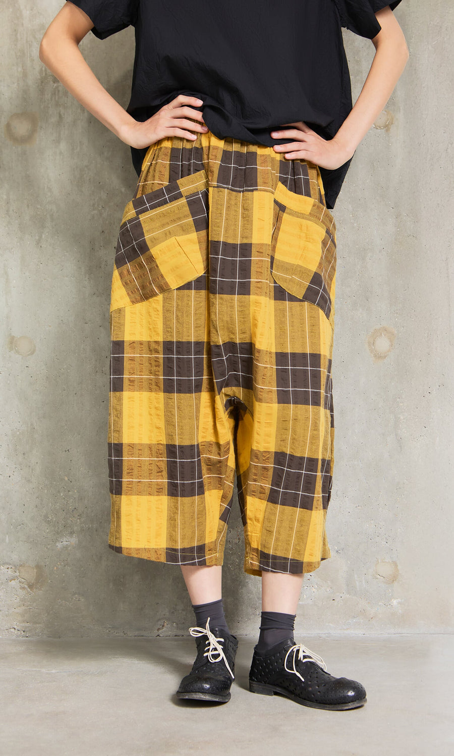 Plümo- Person wears Pimlico Pants in yellow and brown checks, a black shirt, and black shoes by a concrete wall.