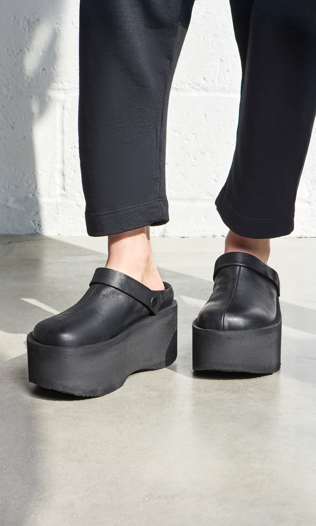 Plümo- Person wearing Kim Clogs—Italian handcrafted black platform clogs—with black pants, standing near a white wall.
