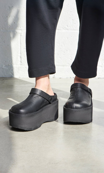 Plümo- Person wearing Kim Clogs—Italian handcrafted black platform clogs—with black pants, standing near a white wall.