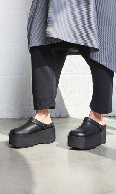 Plümo- Person wearing Kim Clogs, black pants, and a gray skirt near a white brick wall.