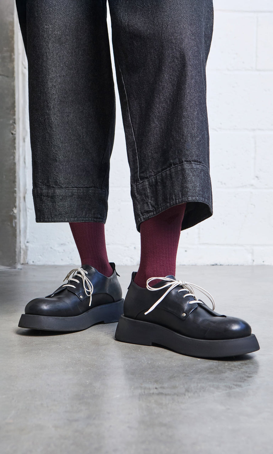 Plümo- Person in wide-legged dark pants, burgundy socks, and Hosier Brogues handcrafted in Italy stands on concrete floor.