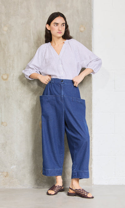 Plümo- Woman stands by a concrete wall in a striped blouse, Polder Pants - Original Denim, and brown sandals.