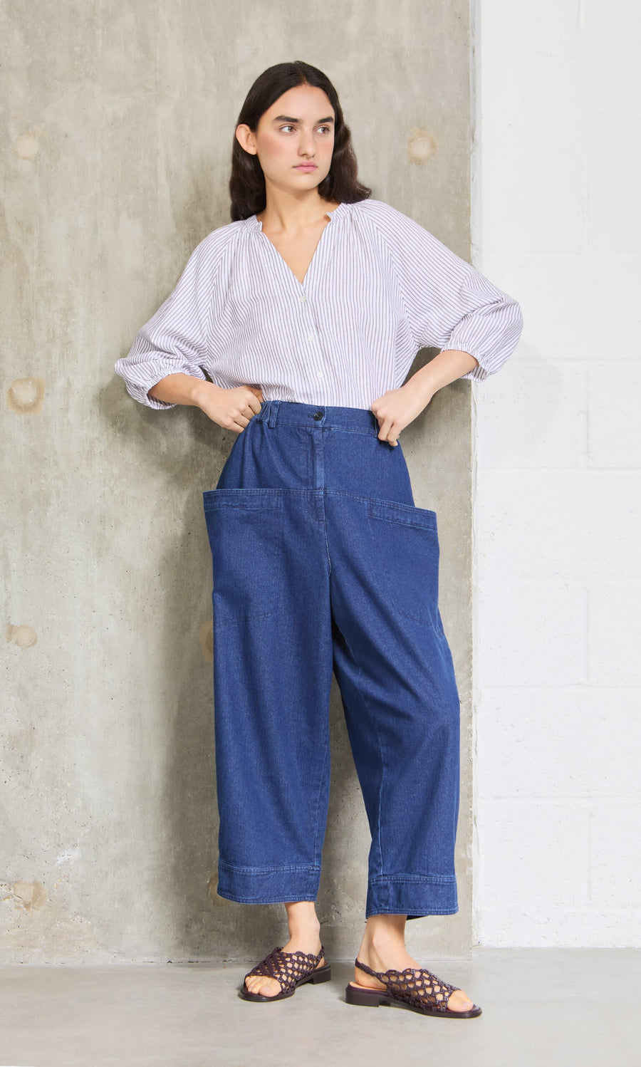 Plümo- Woman stands by a concrete wall in a striped blouse, Polder Pants - Original Denim, and brown sandals.