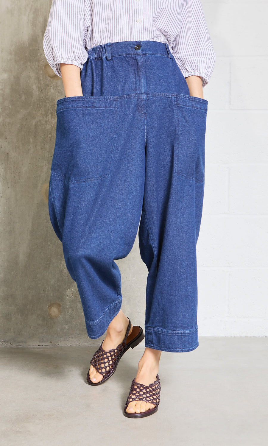 Plümo- Person wearing Polder Pants - Original Denim, a striped top, and brown woven sandals indoors.