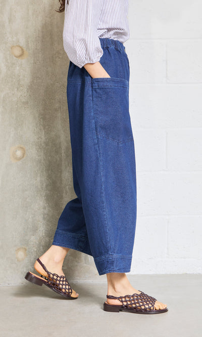 Plümo- Someone wears Polder Pants - Original Denim, featuring an elastic waist, standing indoors by a concrete and white wall.