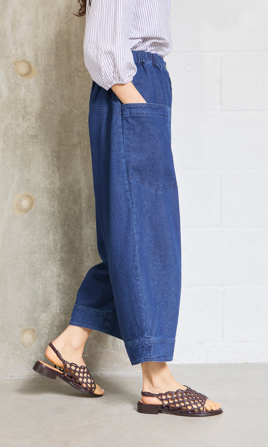 Plümo- Someone wears Polder Pants - Original Denim, featuring an elastic waist, standing indoors by a concrete and white wall.