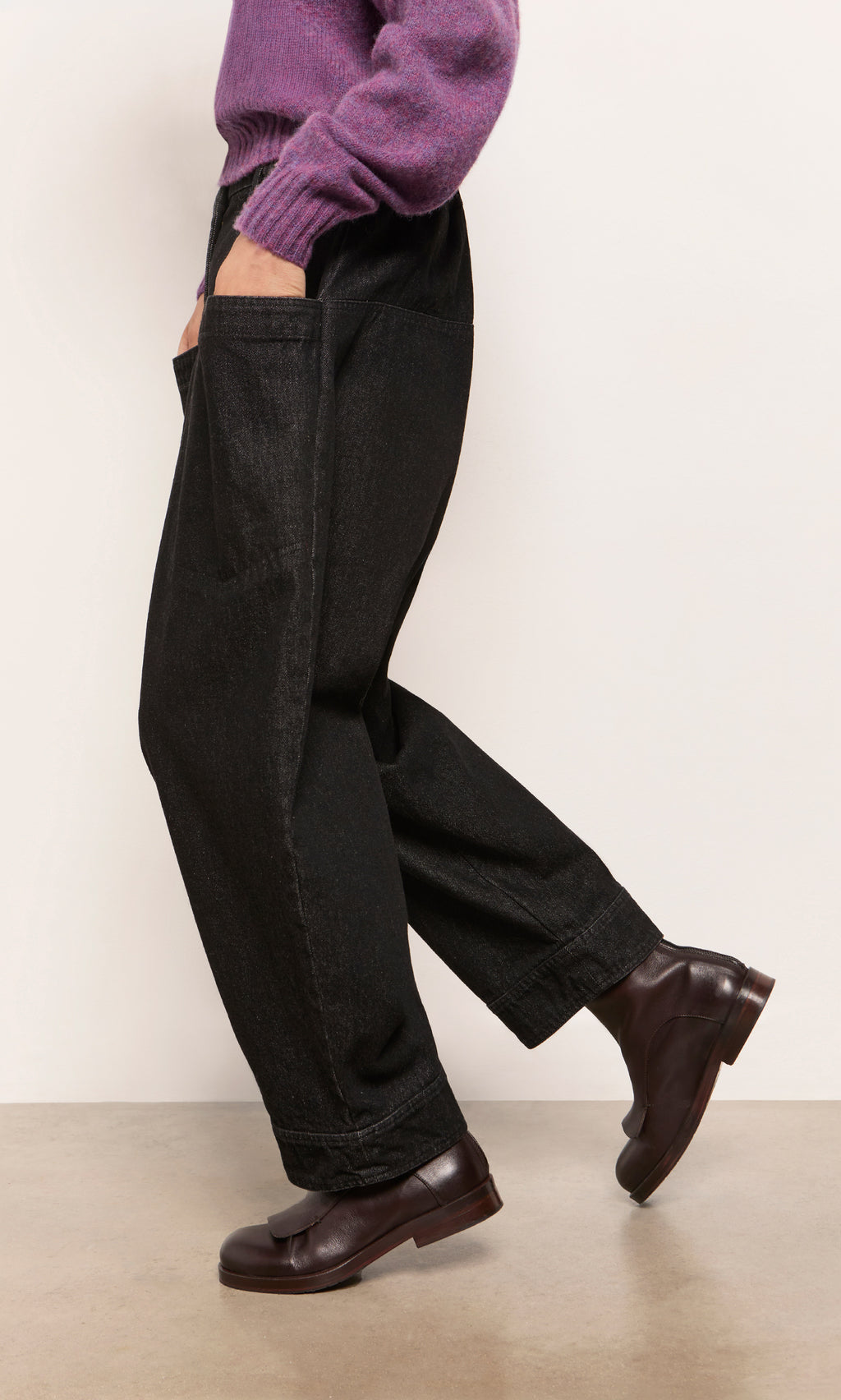 Plümo  Model in Polder Pants - Washed Black, purple sweater, and brown loafers stands by a light wall with one foot raised.