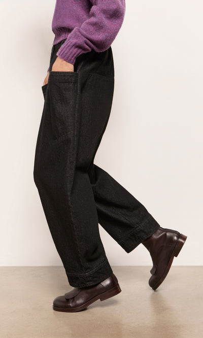 Plümo  Model in Polder Pants - Washed Black, purple sweater, and brown loafers stands by a light wall with one foot raised.