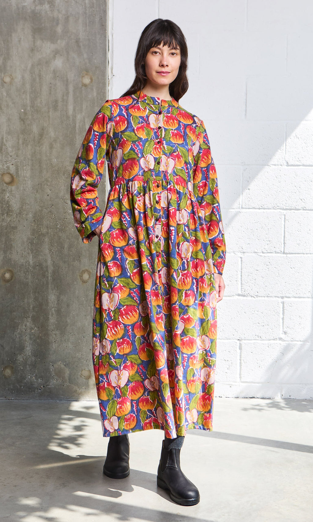 Plümo- Woman in a Braeburn Dress with rosy apple print and black boots stands indoors by a brick and concrete wall.