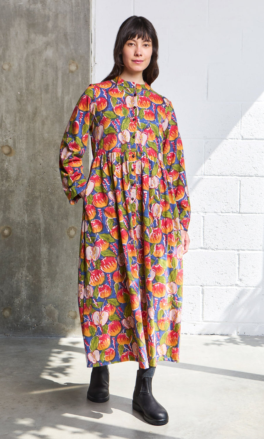 Plümo- Woman in a Braeburn Dress with rosy apple print and black boots stands indoors by a brick and concrete wall.
