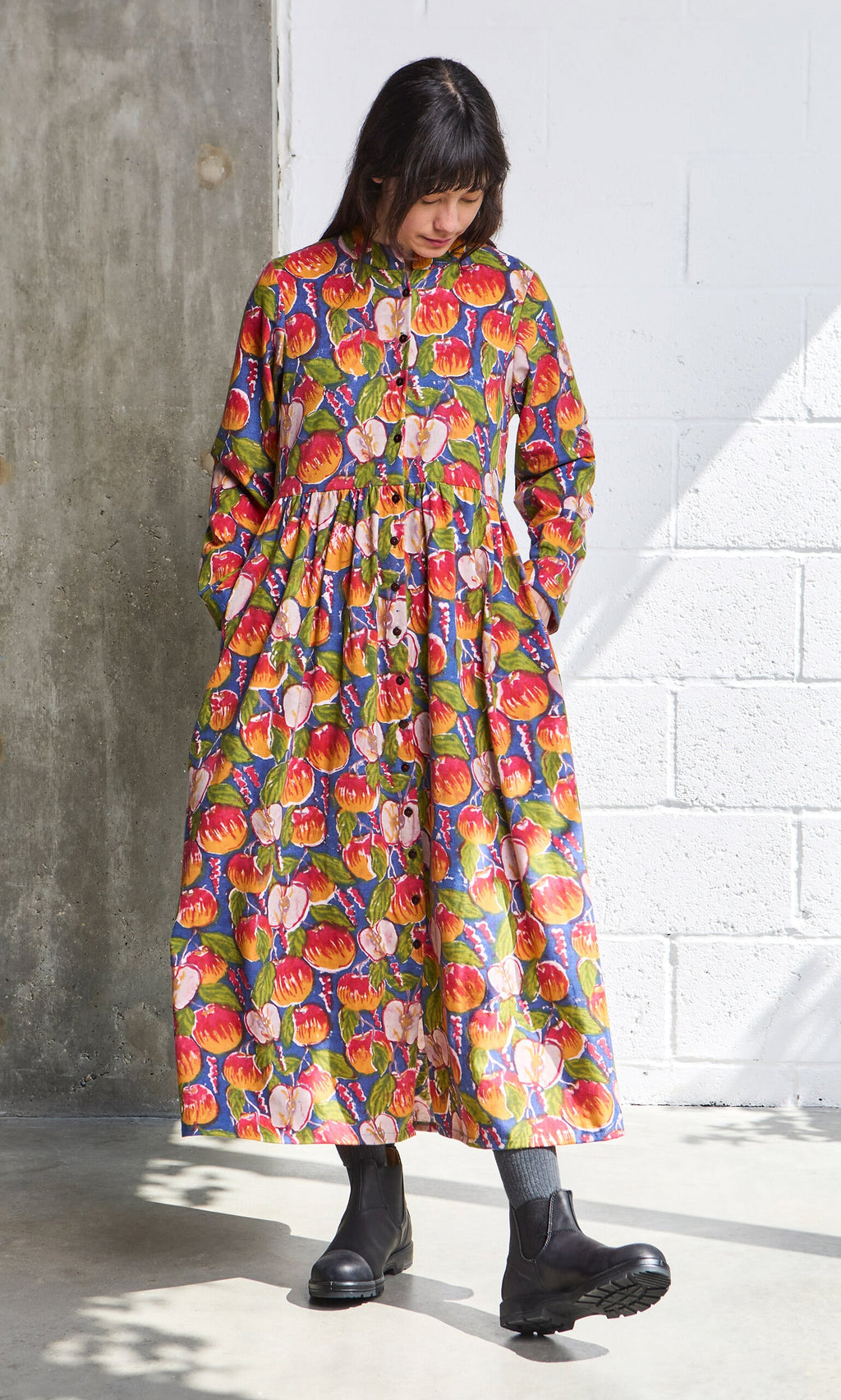 Plümo- Woman in a Braeburn Dress with rosy apple print and pockets, black boots, and gray socks by a concrete white wall.