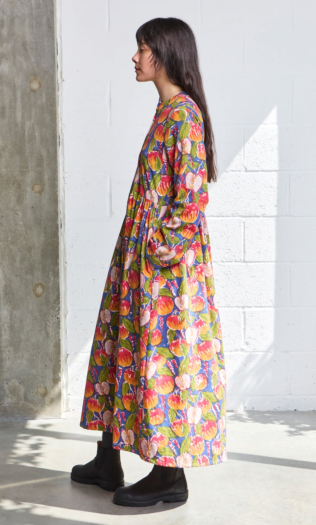 Plümo- Woman in the Braeburn Dress with rosy apple print and black boots stands sideways in a bright, minimalist room.