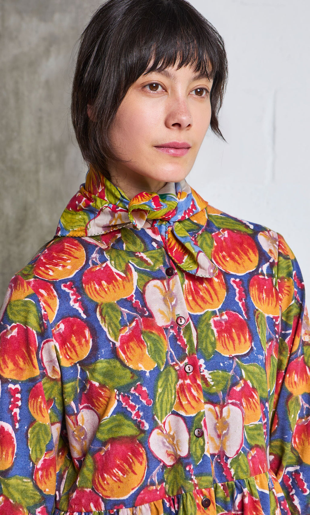 Plümo- Woman with short dark hair in the Braeburn Dress, featuring a rosy apple print and matching neck bow.