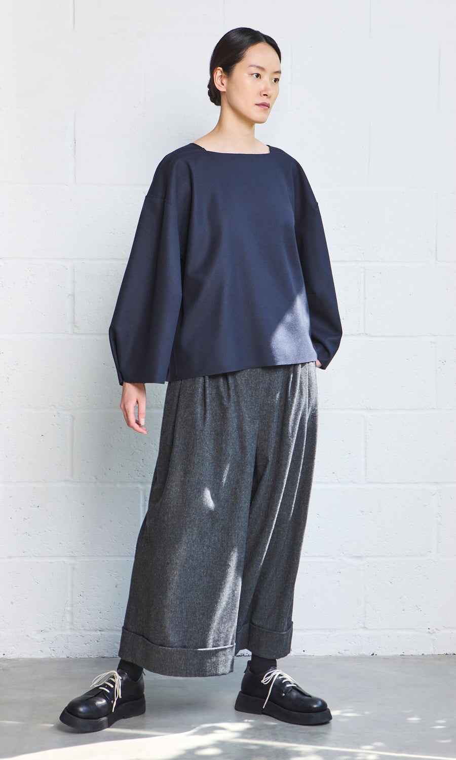 Plümo- Person wearing a loose navy Porter Wool Top, wide gray pants, and black shoes, standing by a white brick wall.