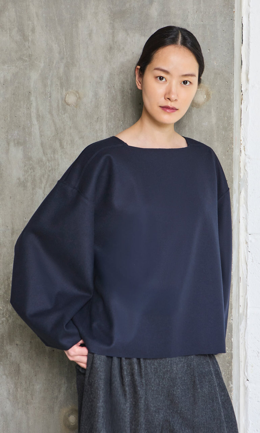 Plümo- Woman with dark hair wears the Porter Wool Top in navy, standing against a textured gray concrete wall, facing the camera.