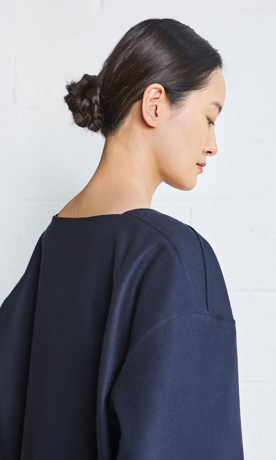 Plümo- Woman with dark hair in a low bun wearing the Porter Wool Top, facing sideways against a white wall background.