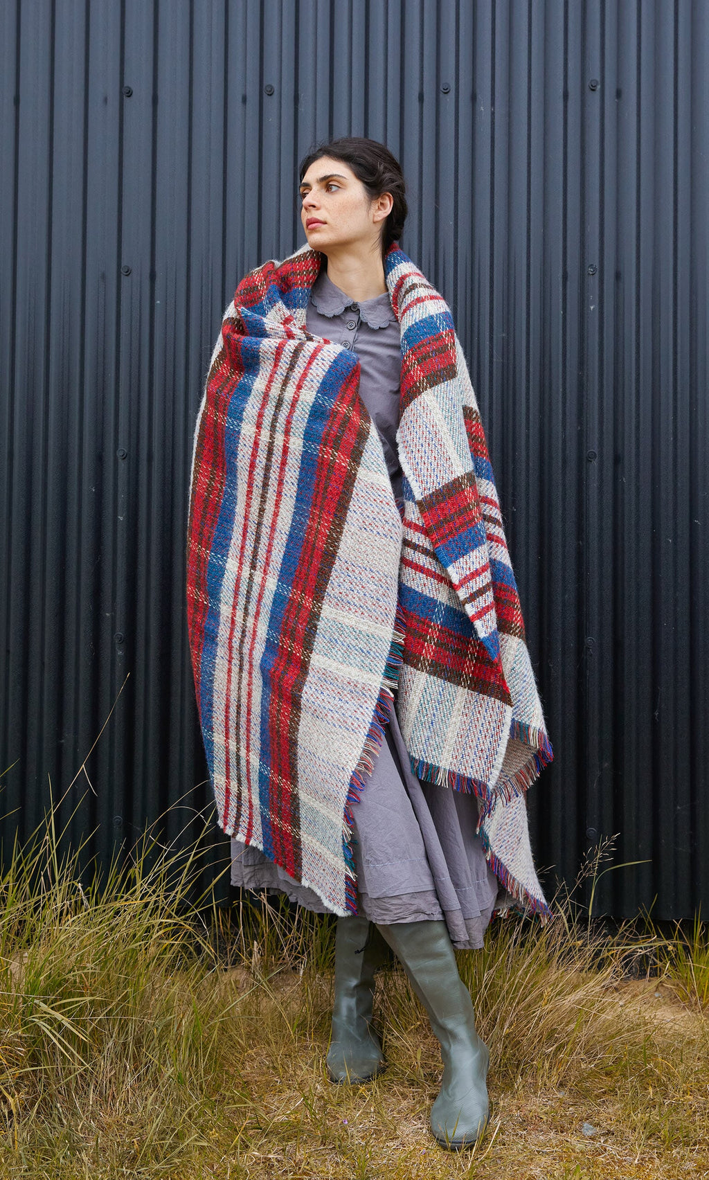 Plümo  A person in a grey dress and boots is wrapped in a Random Throw with a red, blue, and cream plaid pattern.