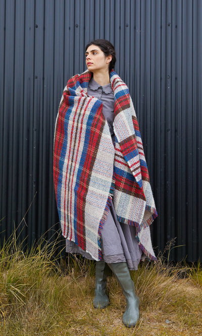 Plümo  A person in a grey dress and boots is wrapped in a Random Throw with a red, blue, and cream plaid pattern.