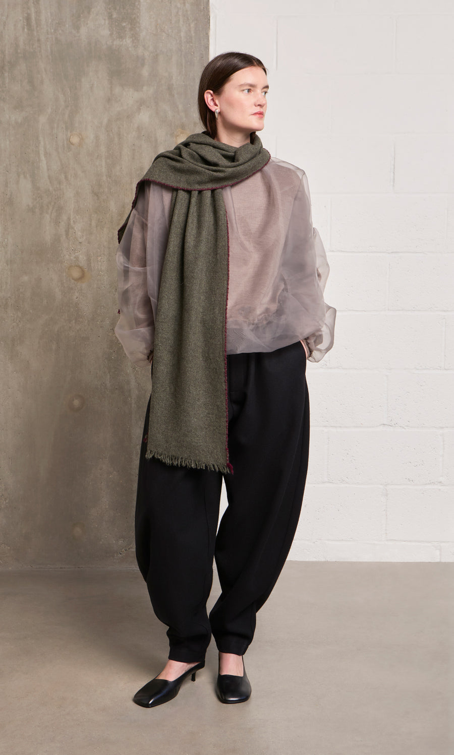 Plümo  Woman in sheer mauve blouse, Cashmere Wrap scarf, black pants, and flats stands by a concrete brick wall.