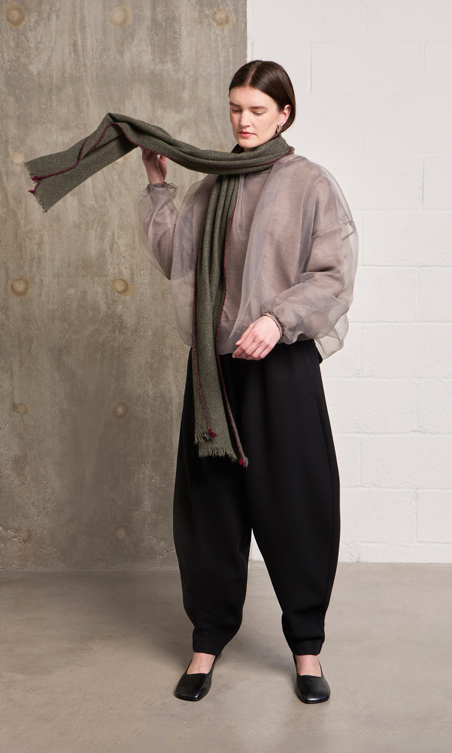 Plümo  Woman in sheer jacket, dark pants, holds a Cashmere Wrap, stands indoors by concrete and white walls.