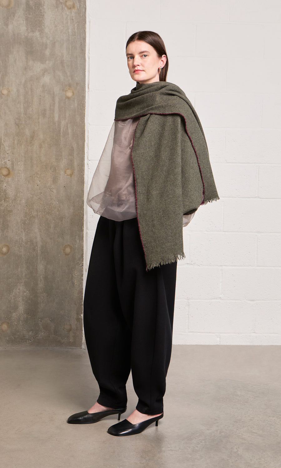 Plümo  Person in a sheer blouse, wide black pants, and a green Cashmere Wrap stands in a minimalist room.