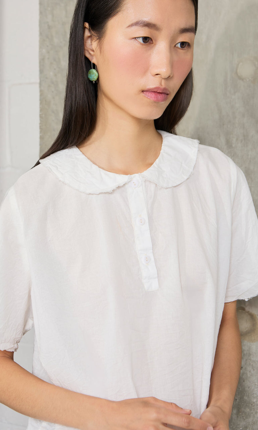 Plümo- A woman in a white blouse and Erika Earrings stands by a concrete wall, looking thoughtful.