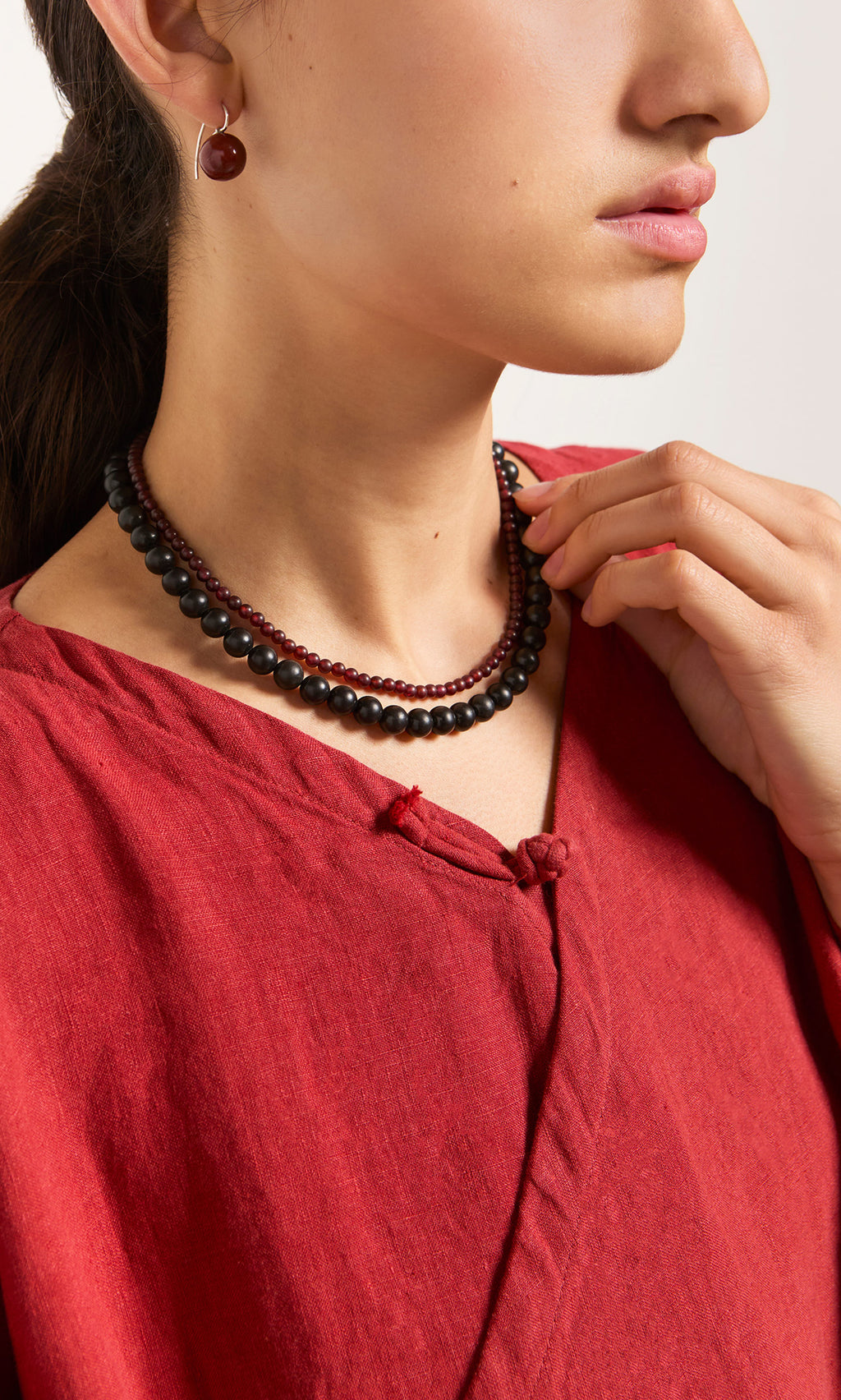 Plümo  Woman in red top models the Rowena Necklace with matching red earrings, hand elegantly touching her necklace.