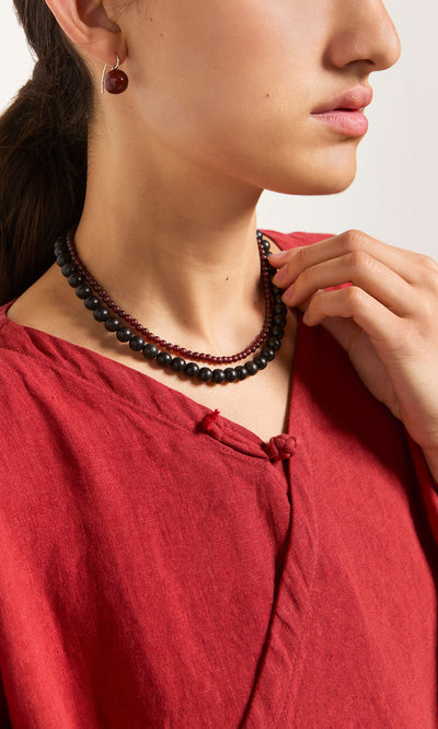 Plümo  Woman in red top models the Rowena Necklace with matching red earrings, hand elegantly touching her necklace.