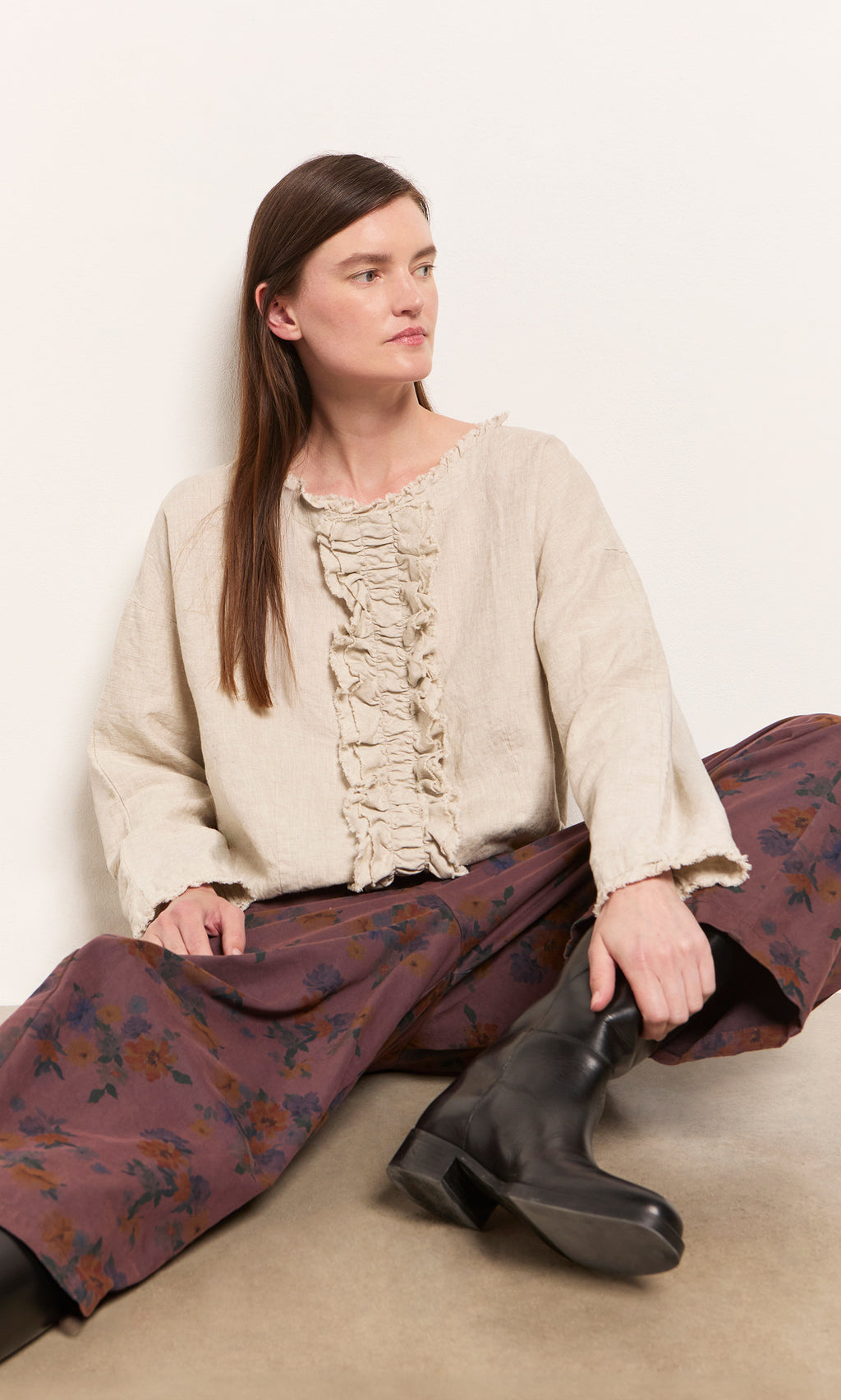 Plümo  Woman seated against a white wall in the Ella Ruffle Top, floral wide-leg pants, and black boots.