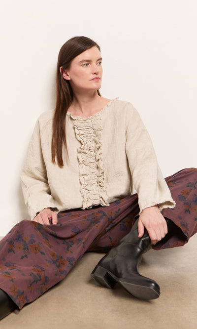 Plümo  Woman seated against a white wall in the Ella Ruffle Top, floral wide-leg pants, and black boots.