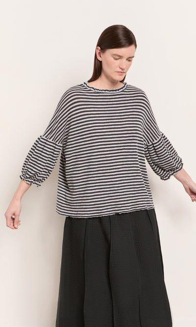 Plümo  Woman wears a Dawn Grey Ruffle Top with balloon sleeves and a long, black textured skirt.