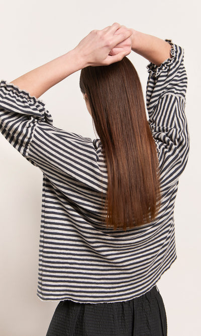 Plümo  Person in a Dawn Grey Ruffle Top, OEKO-TEX certified Danish design, stands with arms raised, back to camera.