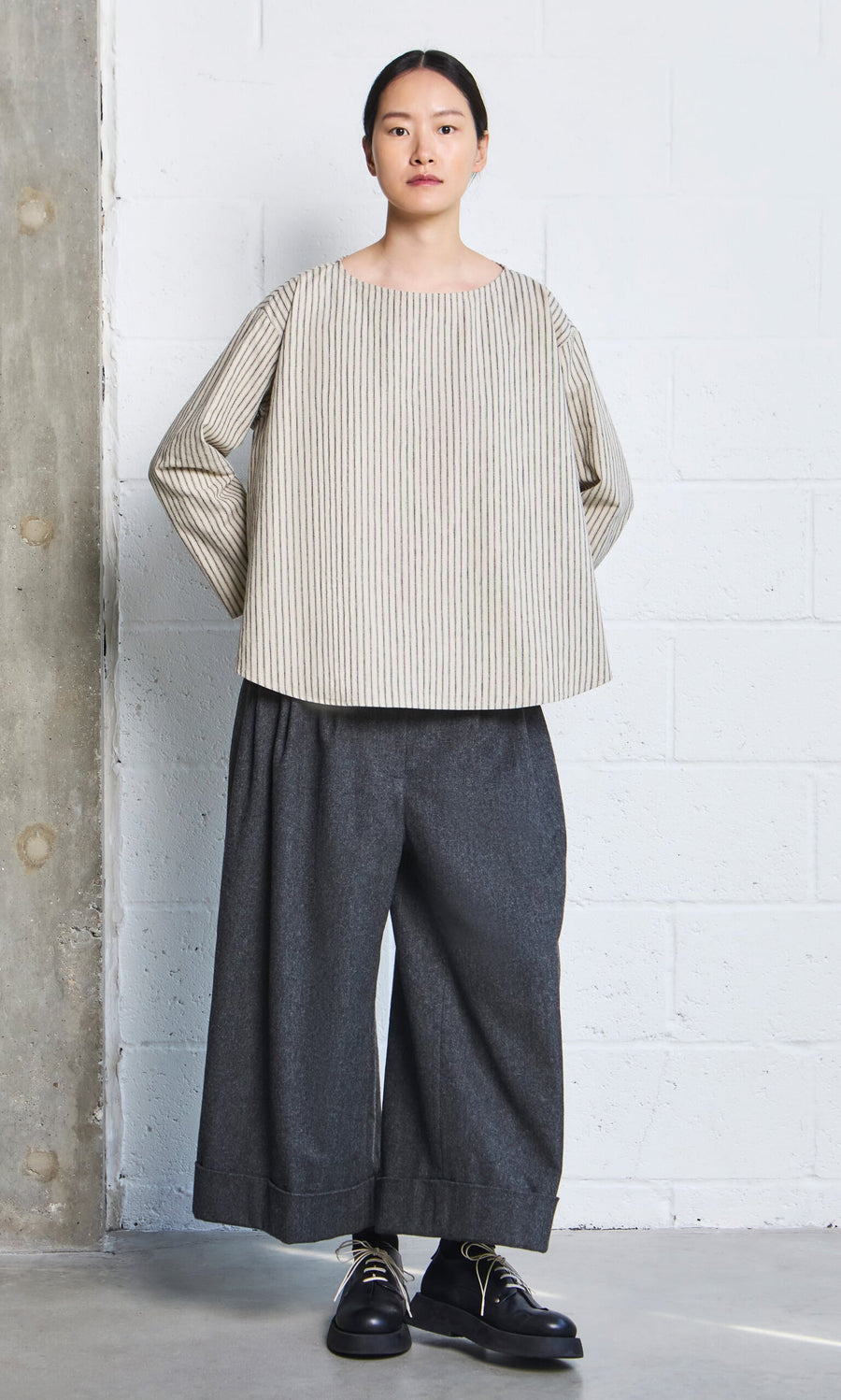 Plümo- Woman wears a Sail Cloth Top and wide-leg gray pants, standing against a white brick wall.