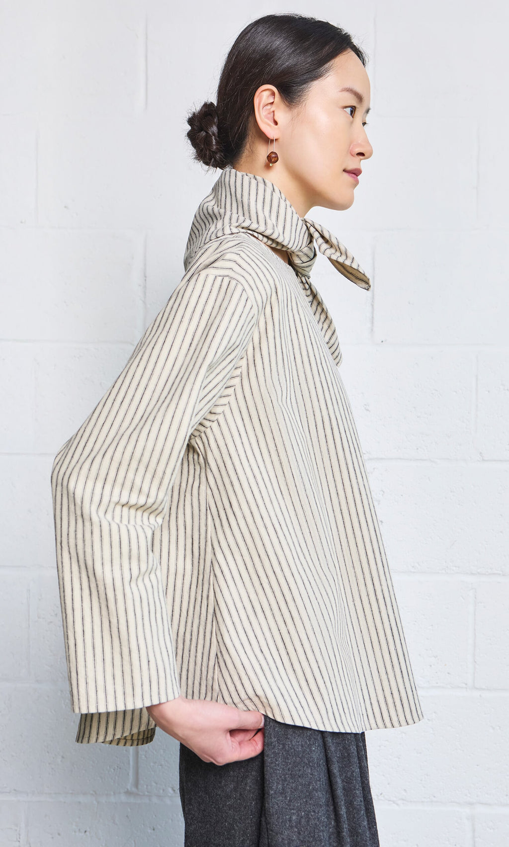 Plümo- Woman wearing the Sail Cloth Top with a high collar, standing sideways against a white wall.