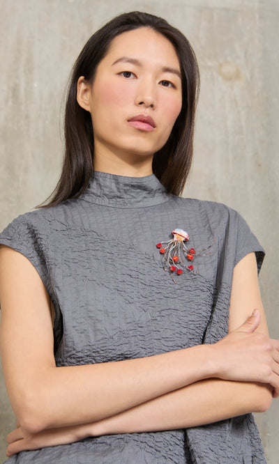 Plümo- Woman in a gray textured top, arms crossed, wears an artisan-made Sea Nettle Brooch, standing against a plain wall.