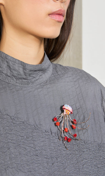 Plümo- A woman in a gray textured top wears the artisan-made Sea Nettle Brooch with red accents on her chest.