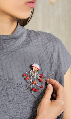 Plümo- A woman wears a textured gray top and a Sea Nettle Brooch with red accents on her chest.