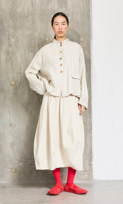 Plümo- A woman in a cream shirt and skirt wears Sharp Red Tights and matching shoes, standing by a concrete wall.
