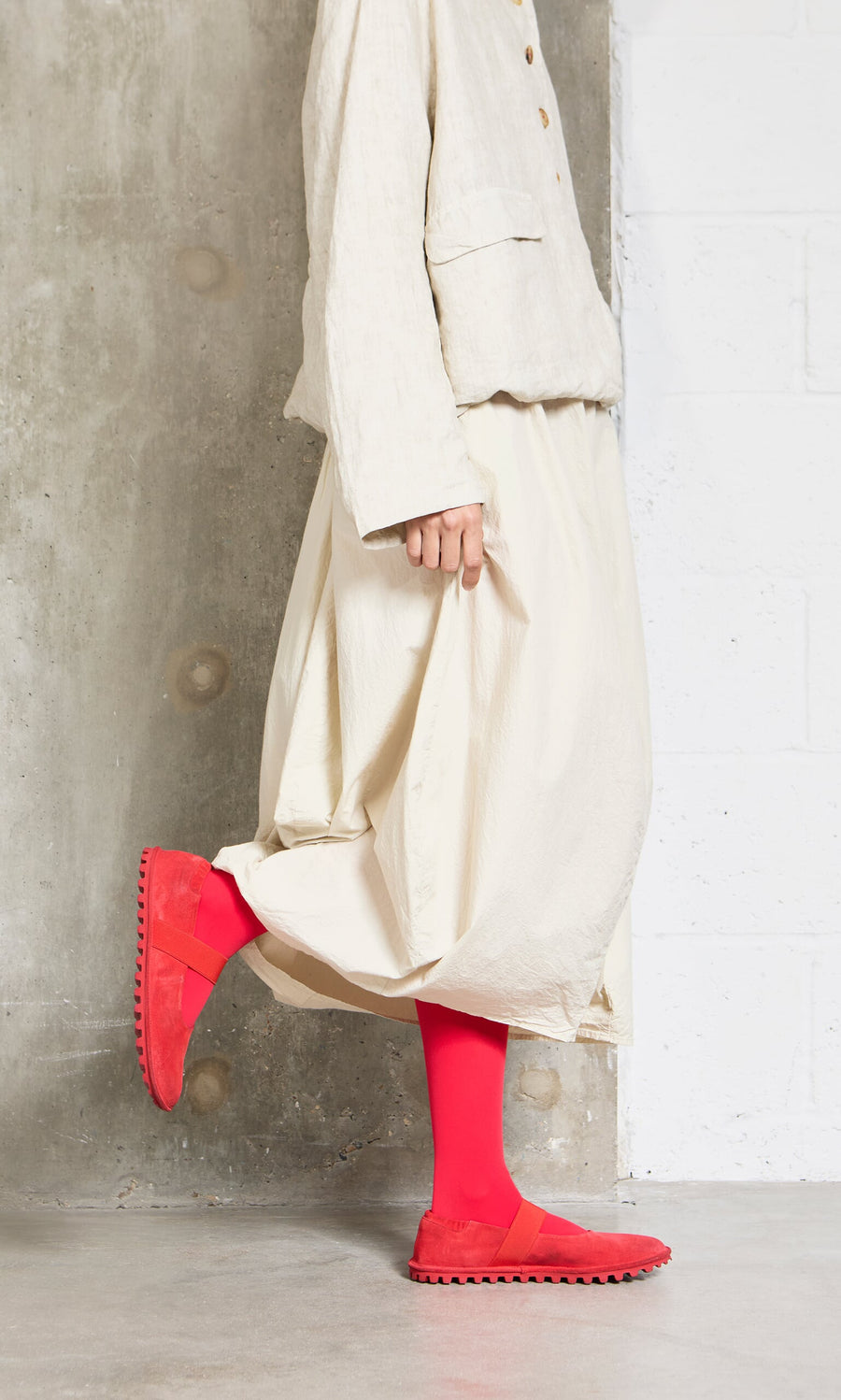 Plümo- Person in loose cream clothing and Sharp Red Tights stands by a concrete wall, holding their skirt hem.