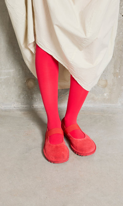 Plümo- Person wears Sharp Red Tights with red Mary Jane shoes and a cream skirt, standing on a concrete floor.